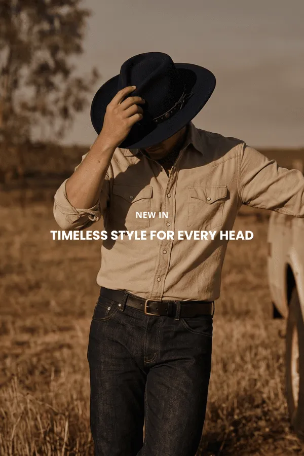 En mand sidder på en trækasse i et studie og bærer en klassisk brun cowboyhat i filt med bred skygge, en mørk denimskjorte og jeans.