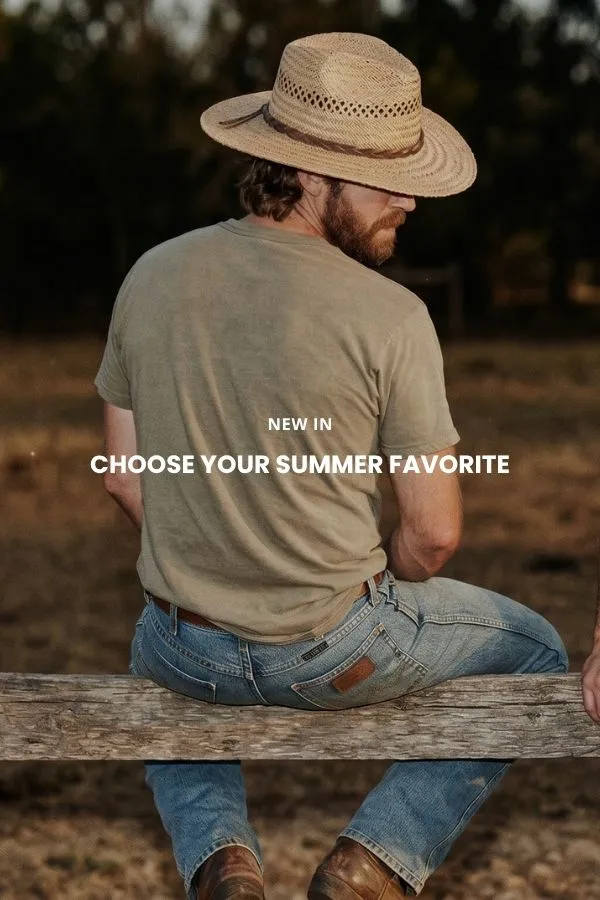 En mand sidder på en trækasse i et studie og bærer en klassisk brun cowboyhat i filt med bred skygge, en mørk denimskjorte og jeans.