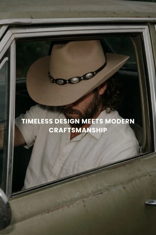 En mand sidder på en trækasse i et studie og bærer en klassisk brun cowboyhat i filt med bred skygge, en mørk denimskjorte og jeans.